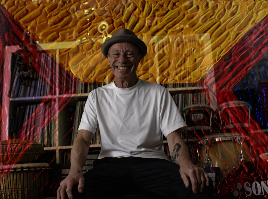 Rich Thair seated in his studio in front of shelves of records and congas, with red and yellow paint-like projections washing over the room.