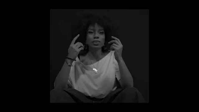 Tia Zakura seated, hands raised, in a black and white video still.